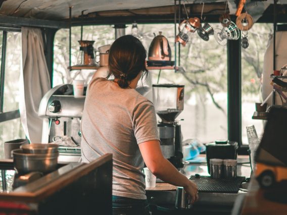 Image shows a young person working in a cafe. Their back is to the camera and they are placing a drink on the counter.