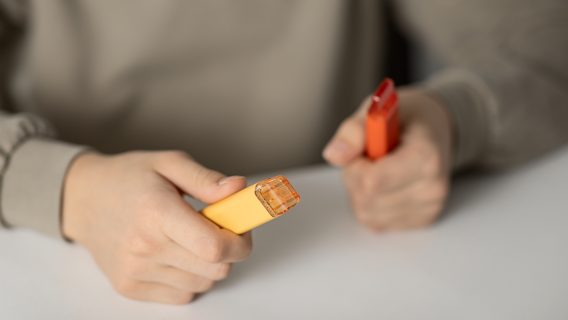 Hands holding one orange and one red vape on a white table